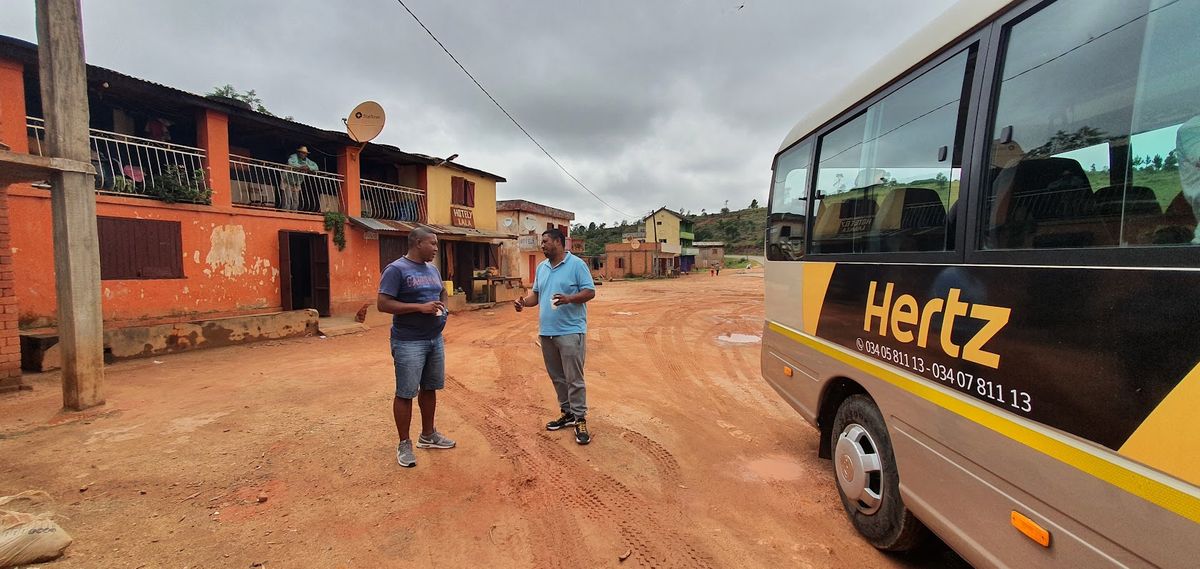 Minibus de location arrêté sur une place de latérite détrempée dans un village des hautes terres malgaches sous un ciel de saison des pluies